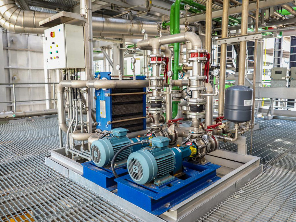Two skid-mounted centrifugal pumps with electric motors, stainless steel pipework, a plate heat exchanger and a control panel on a steel grating floor