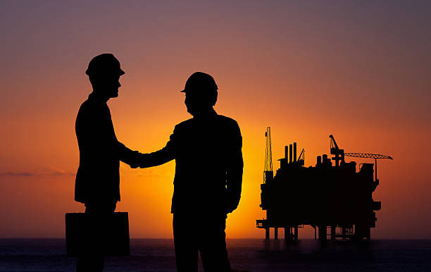 Two people in hard hats shaking hands in silhouette with an offshore production platform at sunset behind them