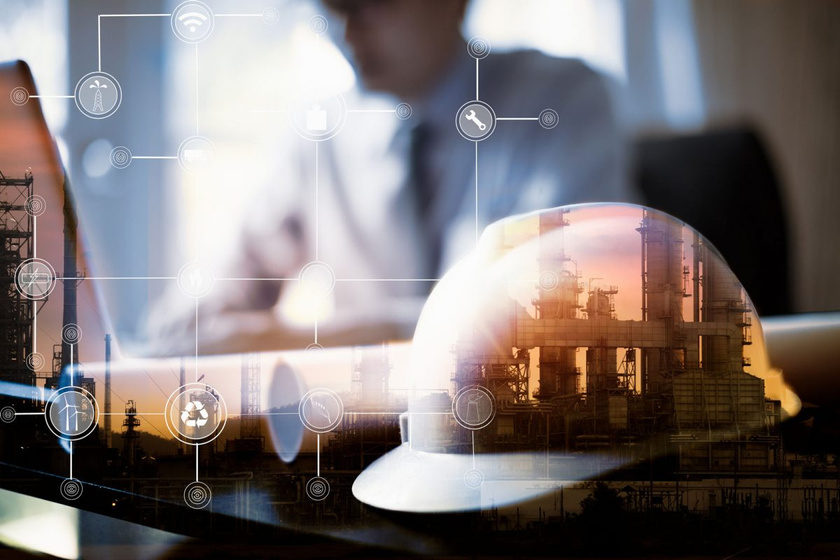 White hard hat resting on a desk in front of an engineer working at a laptop, overlaid with a refinery skyline and industrial icons